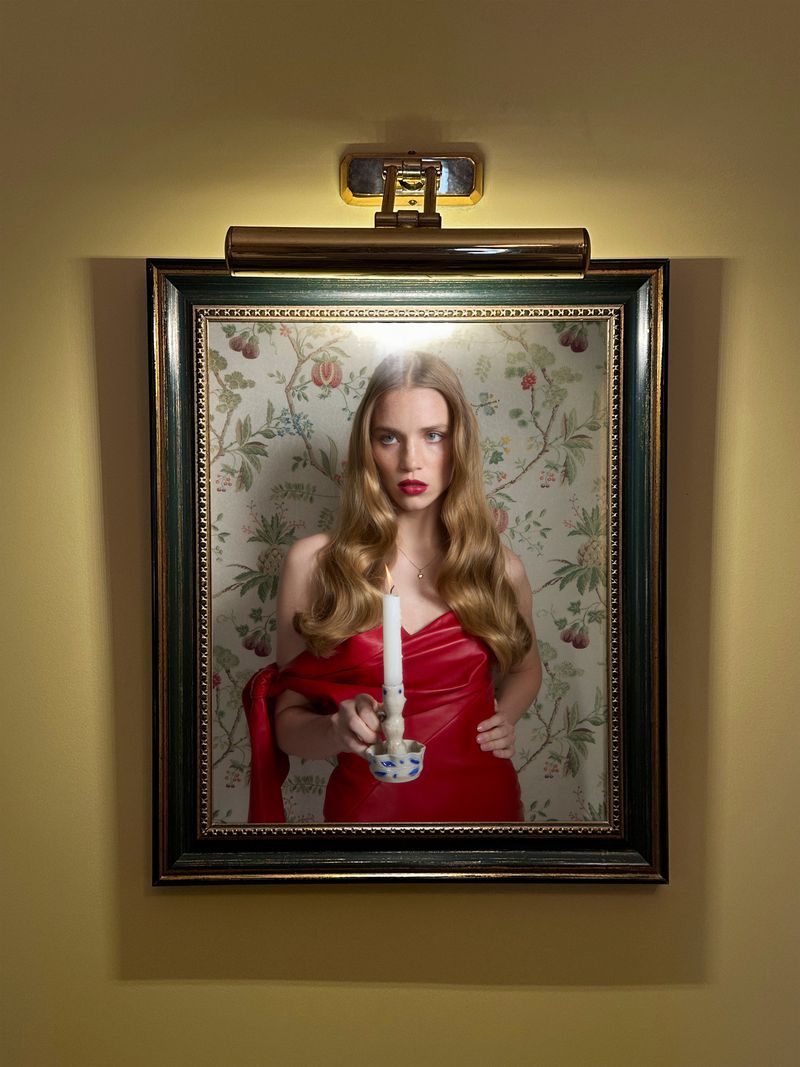 Woman in red dress holding candle in front of floral wallpaper, framed and lit by a picture light on yellow wall. Portrait.