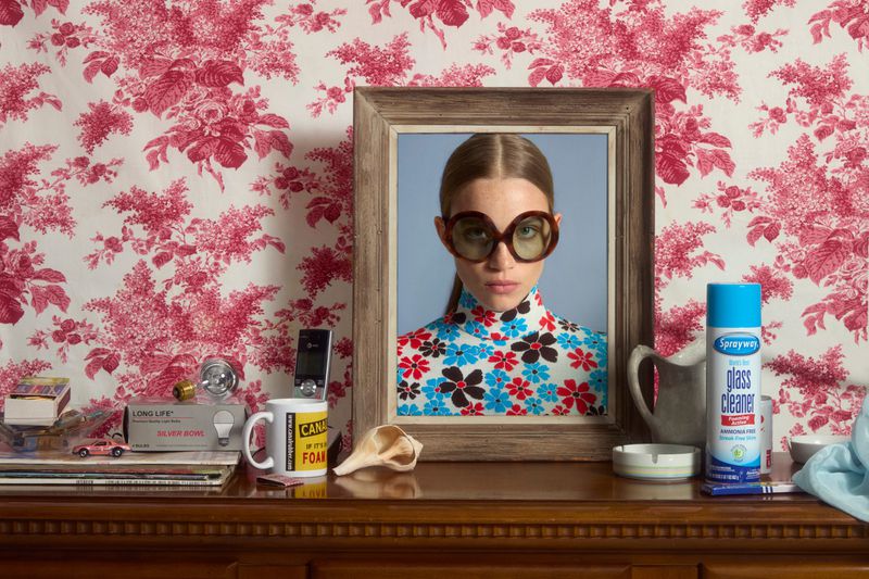 Framed portrait of a woman with round glasses and floral top on a dresser with cleaning supplies against a floral wallpaper background.