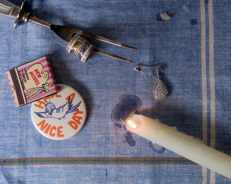 Still life with burning candle, jewelry, matchbook, and button on a blue striped fabric surface, creating an intimate composition.