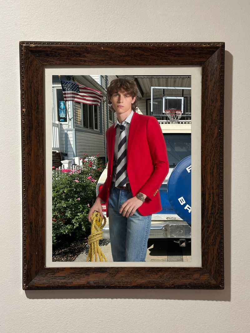 Photo of a young adult male in a red blazer and striped tie standing in front of a vintage vehicle and house with an American flag.