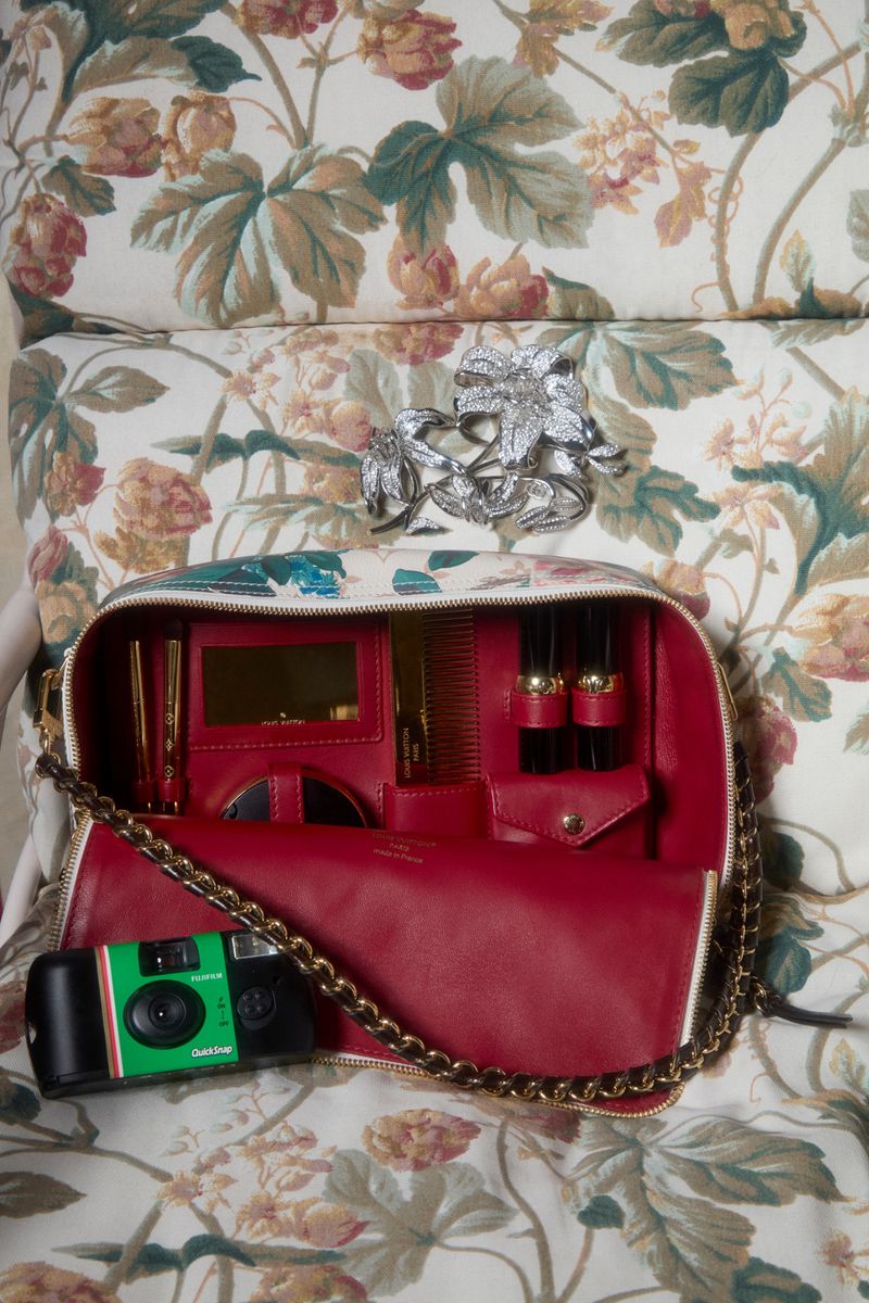 Open makeup bag with lipstick tubes, comb, compact mirror, and disposable camera. Floral pattern wallpaper and leaf-shaped broach.