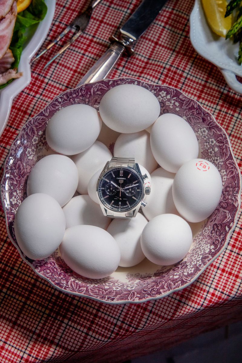 Overhead shot of a stainless steel watch resting on a bowl filled with white eggs, displayed on a red and white plaid surface.