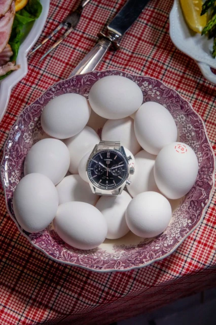Overhead shot of a stainless steel watch resting on a bowl filled with white eggs, displayed on a red and white plaid surface.