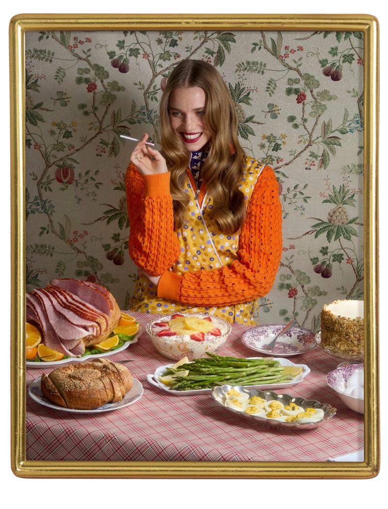 Framed portrait of a woman at a dining table laden with ham, pineapple salad, bread, and deviled eggs, set against floral wallpaper.