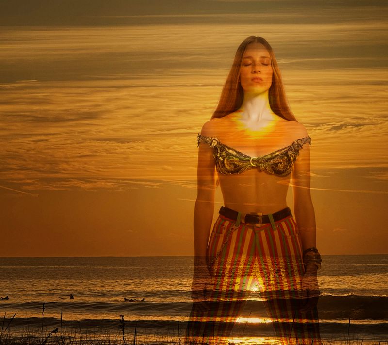 Woman in ornate top and striped pants stands at a shoreline at sunset, gazing forward in a double exposure image.