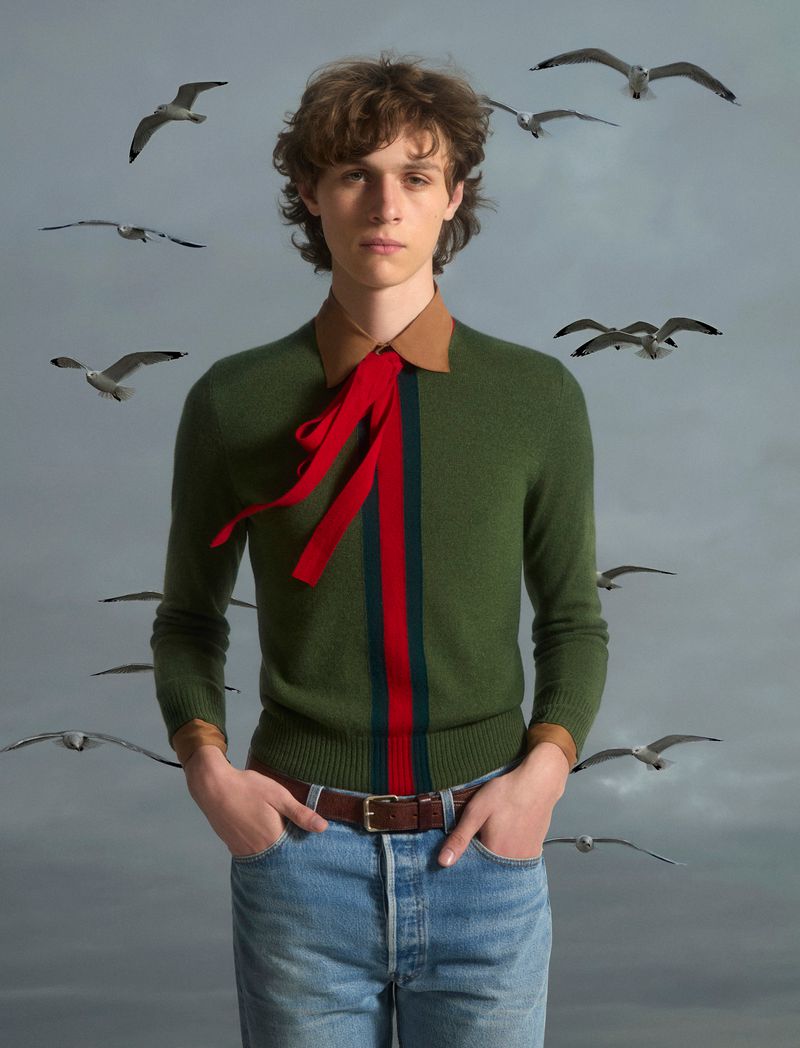 Portrait of a curly-haired male model in a green sweater and jeans, surrounded by seagulls on a cloudy day for a fashion editorial.