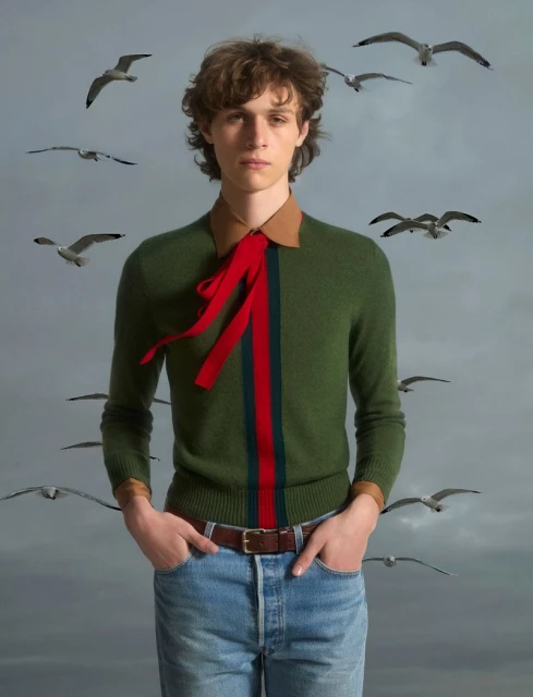 Portrait of a curly-haired male model in a green sweater and jeans, surrounded by seagulls on a cloudy day for a fashion editorial.