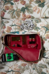 An open floral vanity bag with red interior holding cosmetics, beside a disposable camera on floral upholstery.