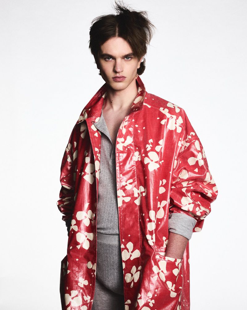 A young adult man poses in a vibrant red floral-patterned overcoat and gray top against a bright white backdrop in a fashion shot.