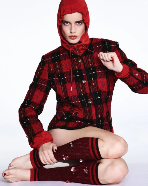 A fashion shot of a woman in a red hooded jacket and leg warmers posing against a white background in a photo studio, full studio lighting.