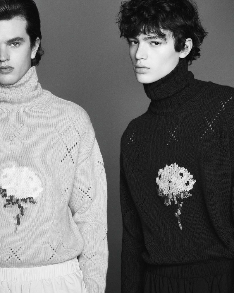 Two young men pose in a studio wearing knit turtlenecks with floral designs. A grayscale studio shot with neutral background.
