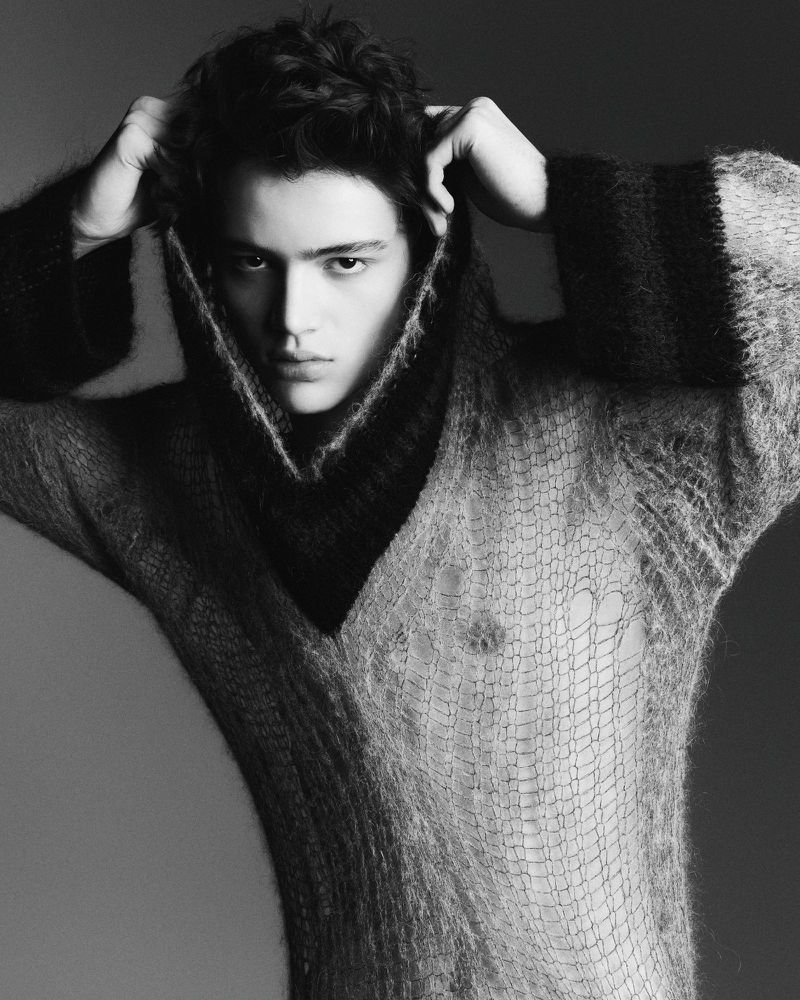 Portrait of a young adult man wearing a dark and light knit sweater, looking directly at the camera in a studio setting.