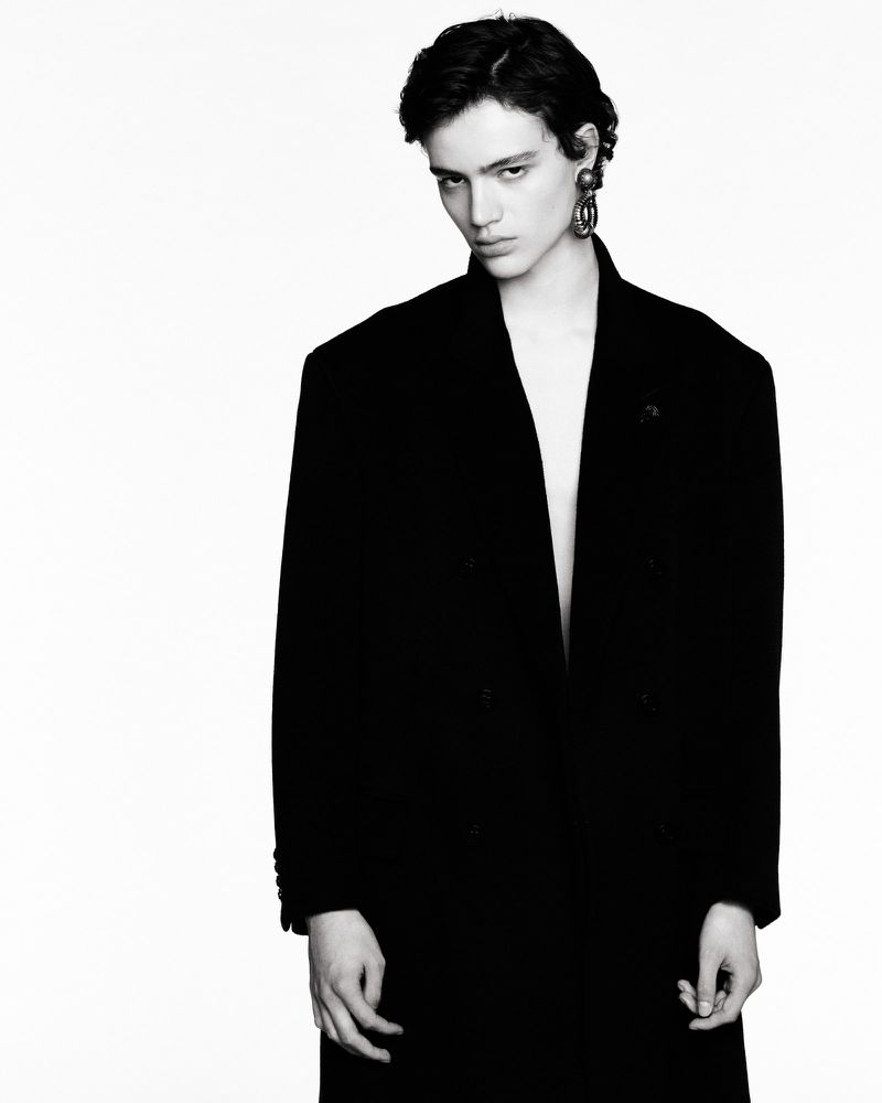 Monochrome portrait of a person with short dark hair wearing an open black coat and large earrings against a stark white background.