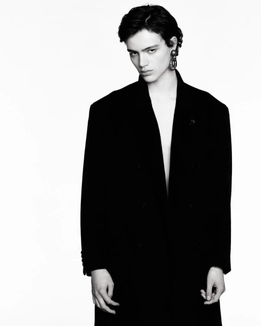 Monochrome portrait of a person with short dark hair wearing an open black coat and large earrings against a stark white background.