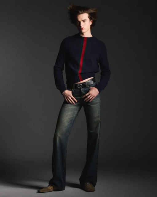 Portrait of a young adult male fashion model in a navy blue sweater and flare jeans posing against a gray studio background.