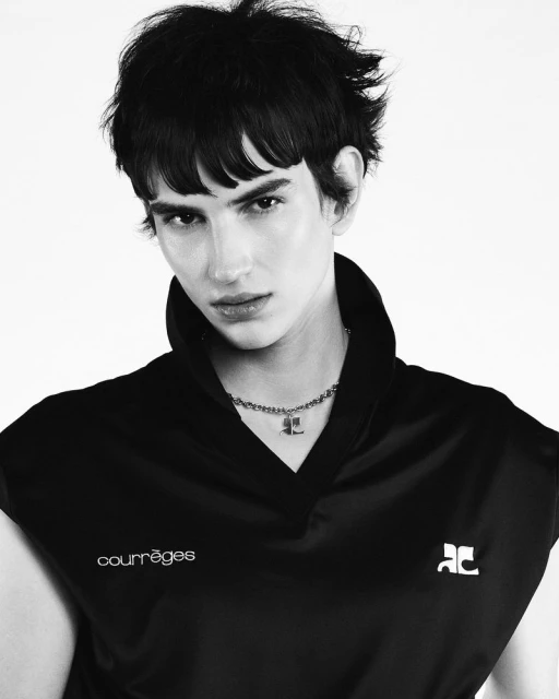 Monochrome portrait of a young adult male with short dark hair, wearing a black Courrèges sleeveless shirt against a white backdrop.