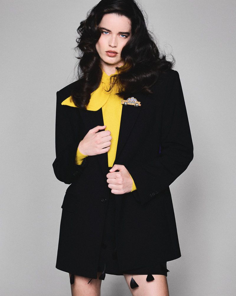 Brunette woman in black blazer and bright yellow top poses in a studio environment with soft lighting for a fashion editorial.