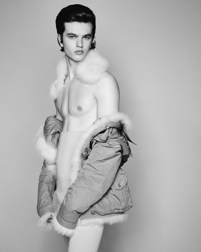Black and white portrait of shirtless man wearing fur-trimmed parka jacket looking at the camera in a photo studio setting.