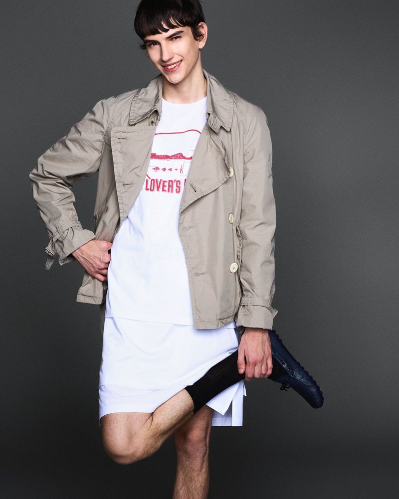 Portrait of a young adult male in a neutral trench coat and white t-shirt, holding a shoe, in a studio with a grey backdrop.