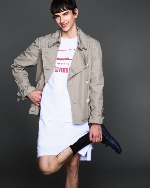 Portrait of a young adult male in a neutral trench coat and white t-shirt, holding a shoe, in a studio with a grey backdrop.