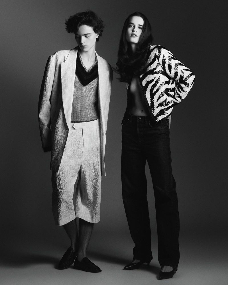 Monochrome fashion shot of a young adult man and woman posing in a studio, wearing stylish patterned blazers and relaxed-fit pants.