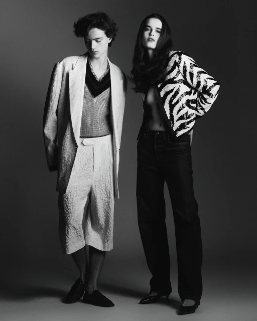 Monochrome fashion shot of a young adult man and woman posing in a studio, wearing stylish patterned blazers and relaxed-fit pants.
