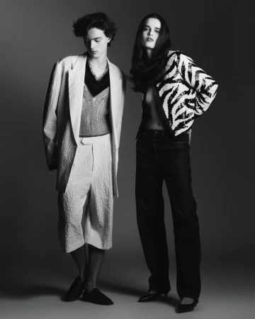 Monochrome fashion shot of a young adult man and woman posing in a studio, wearing stylish patterned blazers and relaxed-fit pants.