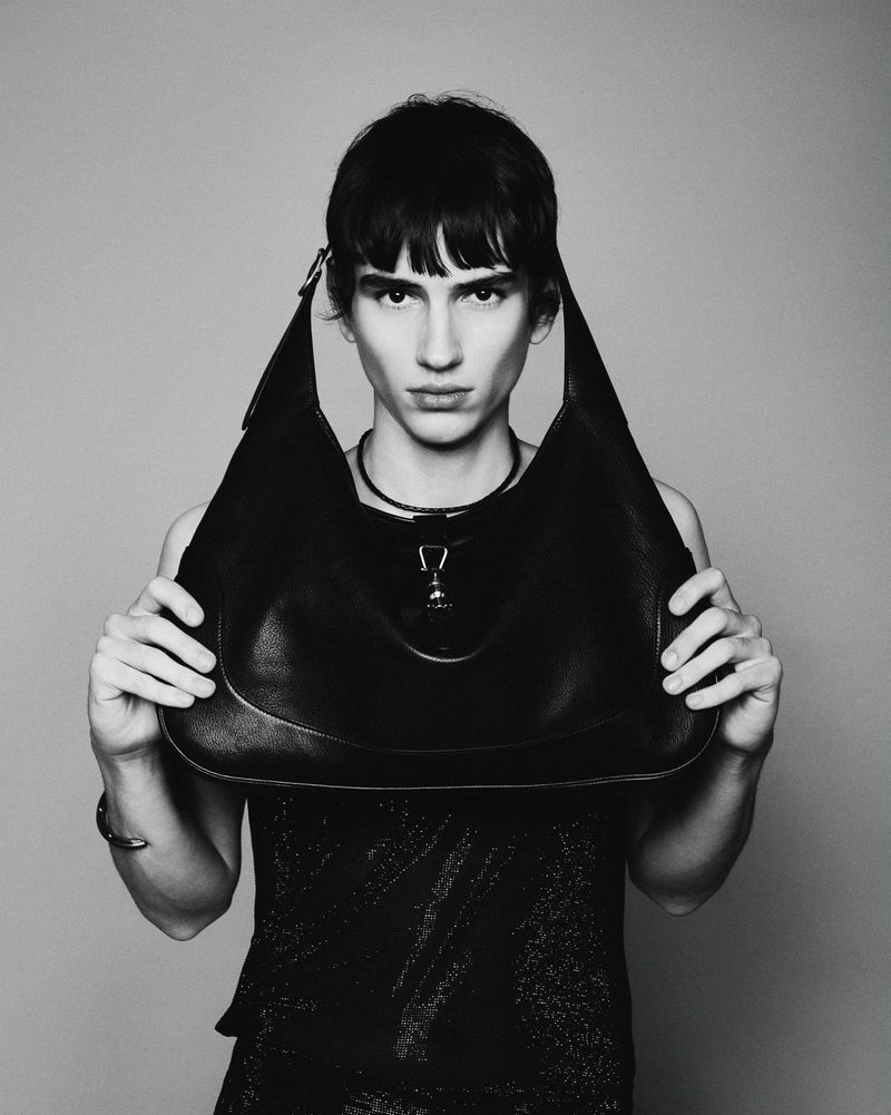 Monochrome portrait of a person holding a black leather hobo bag in a studio setting with a plain backdrop and soft, diffused lighting.
