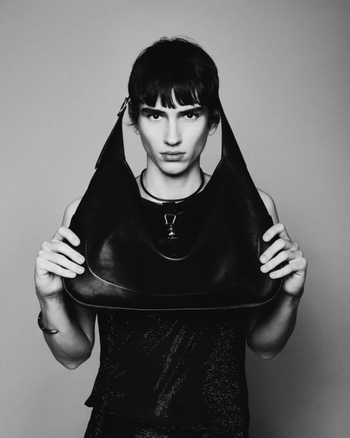 Monochrome portrait of a person holding a black leather hobo bag in a studio setting with a plain backdrop and soft, diffused lighting.