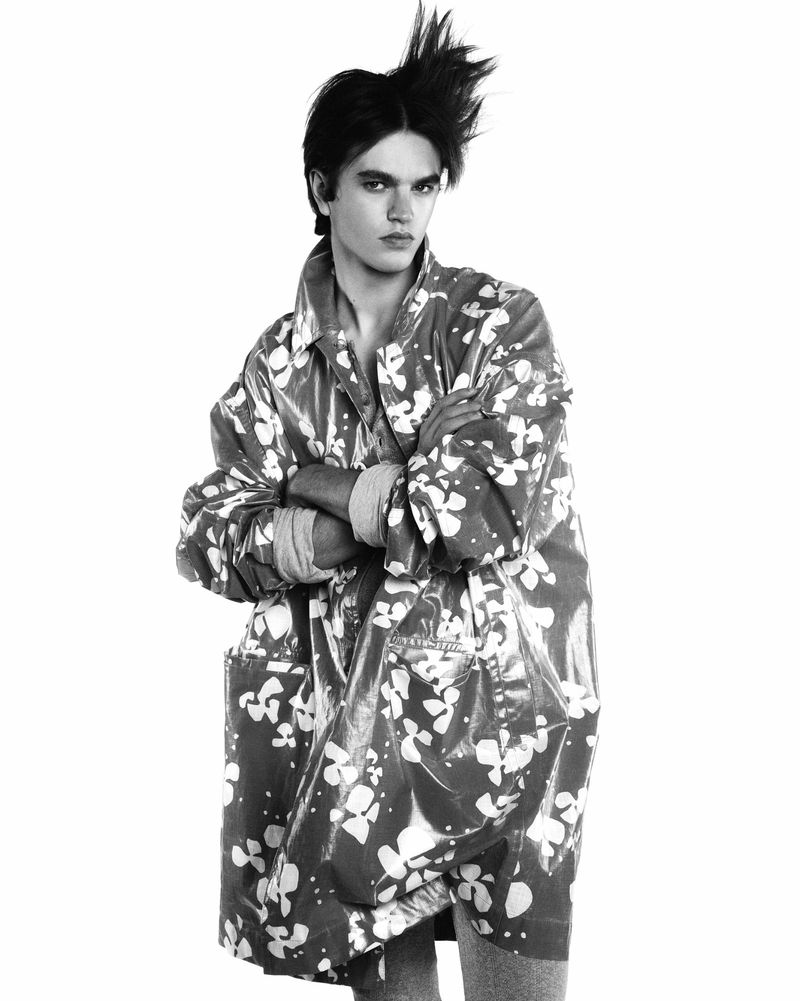 Monochrome portrait of a young adult male with arms crossed wearing a floral print raincoat against a white background in a studio.