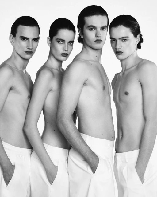 Black and white fashion shot of four people in white pants, standing together closely in a bright, minimalist studio environment.