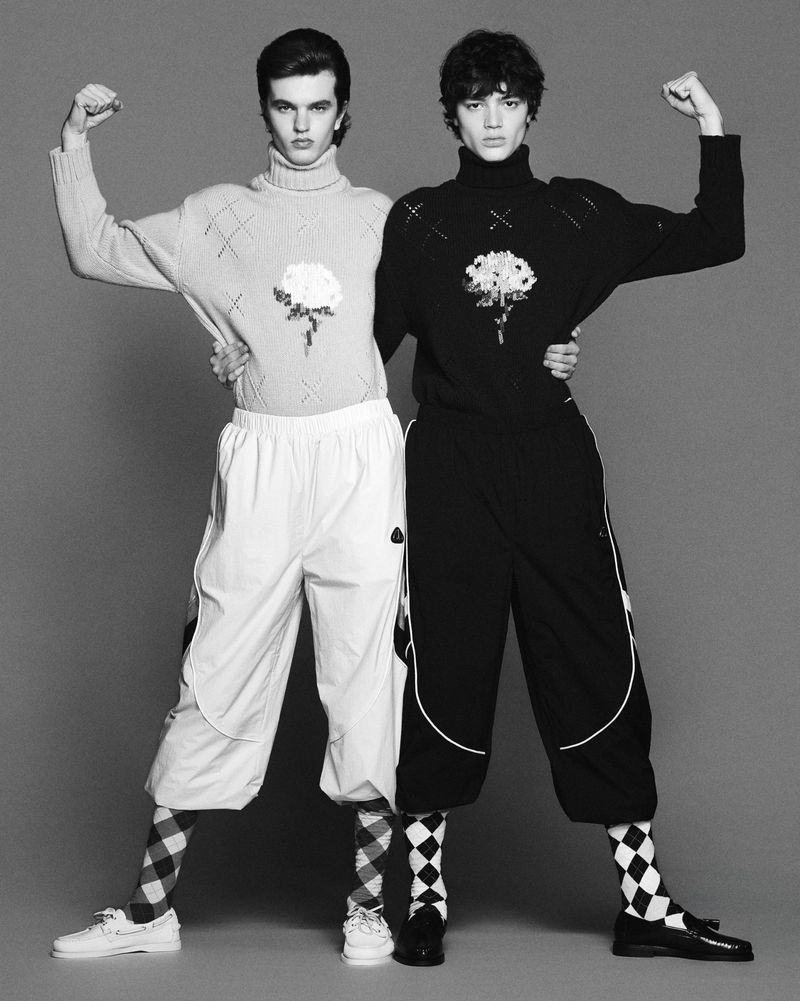 Two male models posing symmetrically in contrasting light and dark knitwear and cropped pants against a grey backdrop.
