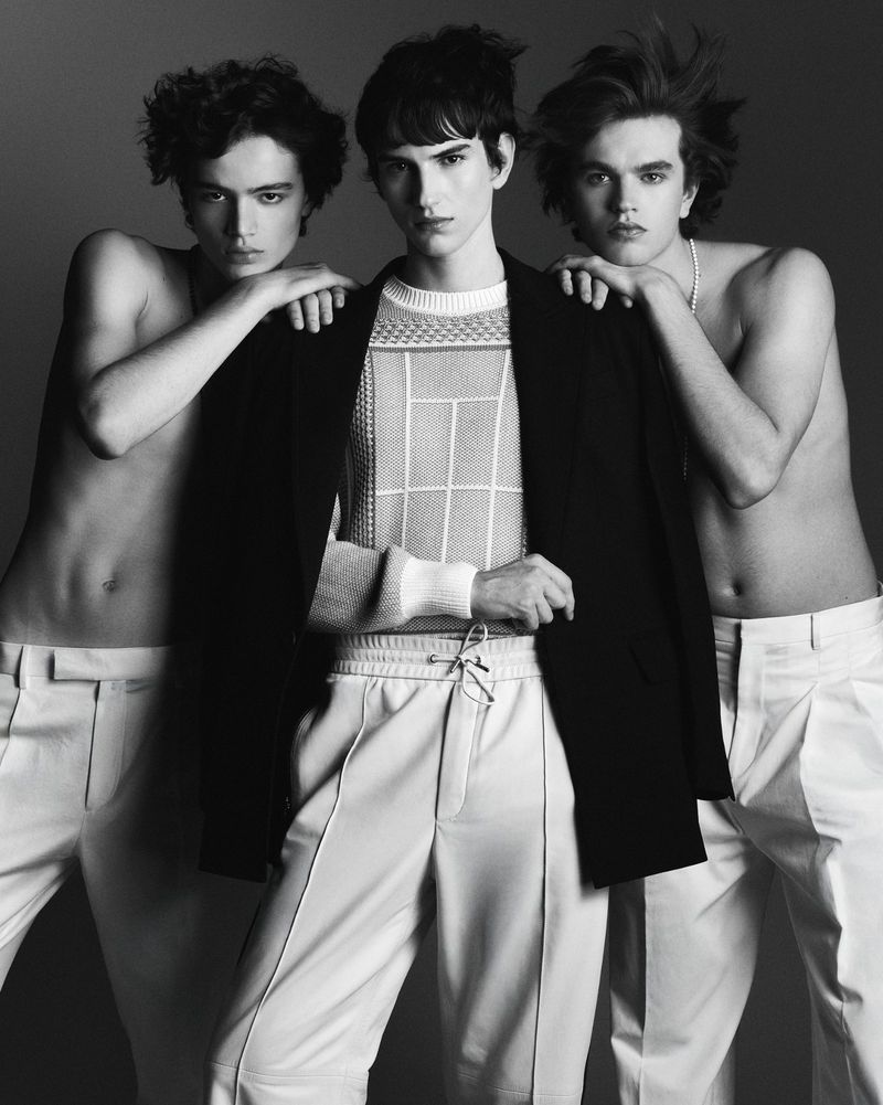 Three young men in a studio shot. The central figure wears a black jacket and patterned knit. The others are shirtless, in white trousers.