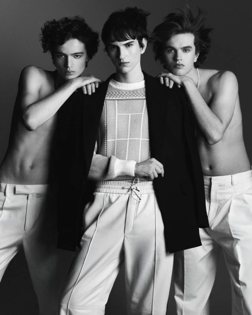 Three young men in a studio shot. The central figure wears a black jacket and patterned knit. The others are shirtless, in white trousers.