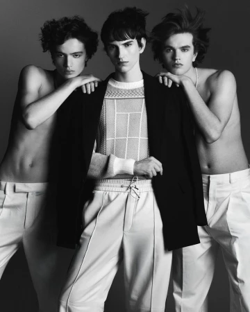 Three young men in a studio shot. The central figure wears a black jacket and patterned knit. The others are shirtless, in white trousers.