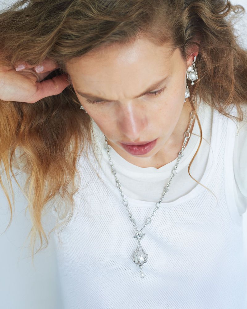 Elegant portrait of an adult woman with long wavy hair wearing a white top and silver drop earrings and necklace in a minimalist setting.