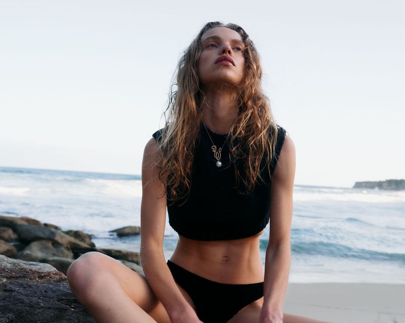Woman in black swimsuit sitting on rocks at beach, gazing upward. Ocean waves in background under a bright sky with distant coastline.