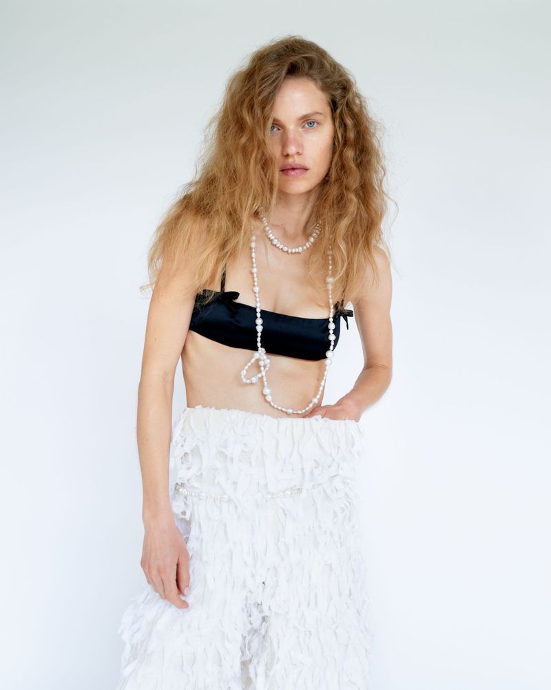 Woman with long wavy hair in a black top and a white skirt with tassels, posing in front of a white background, looking at the camera.