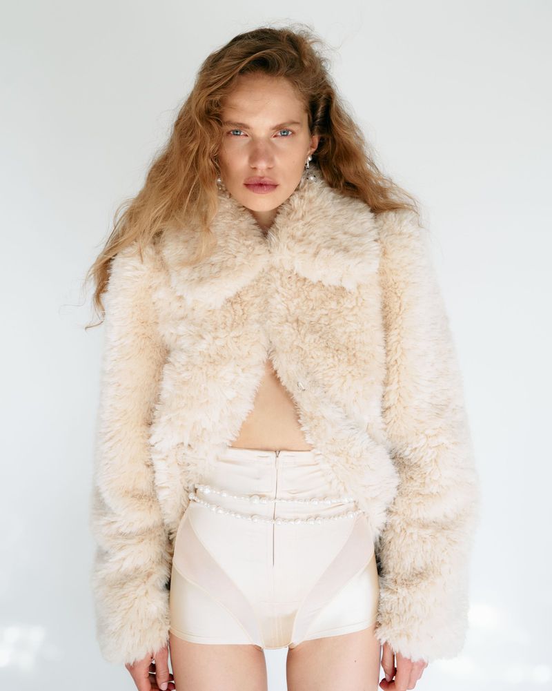 Adult woman poses in a studio wearing a cream-colored, plush jacket and shorts with long blond hair and statement earrings.