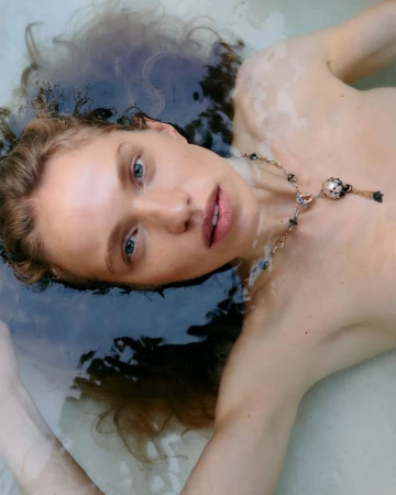 Close-up of a light-skinned adult woman with blue eyes in water, wearing a beaded necklace, looking at the camera with a serene expression.