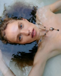 Model Sabine Glud floating in clear shallow water wearing an ornate pendant necklace.