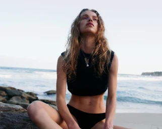 Model Sabine Glud seated on coastal rocks wearing a black sleeveless crop top and bikini bottoms against ocean waves.