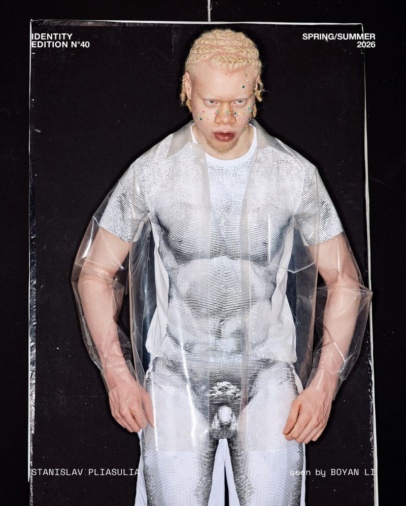 Albino male model poses in a transparent plastic shirt over a graphic shirt. The image is a fashion editorial with a black background.