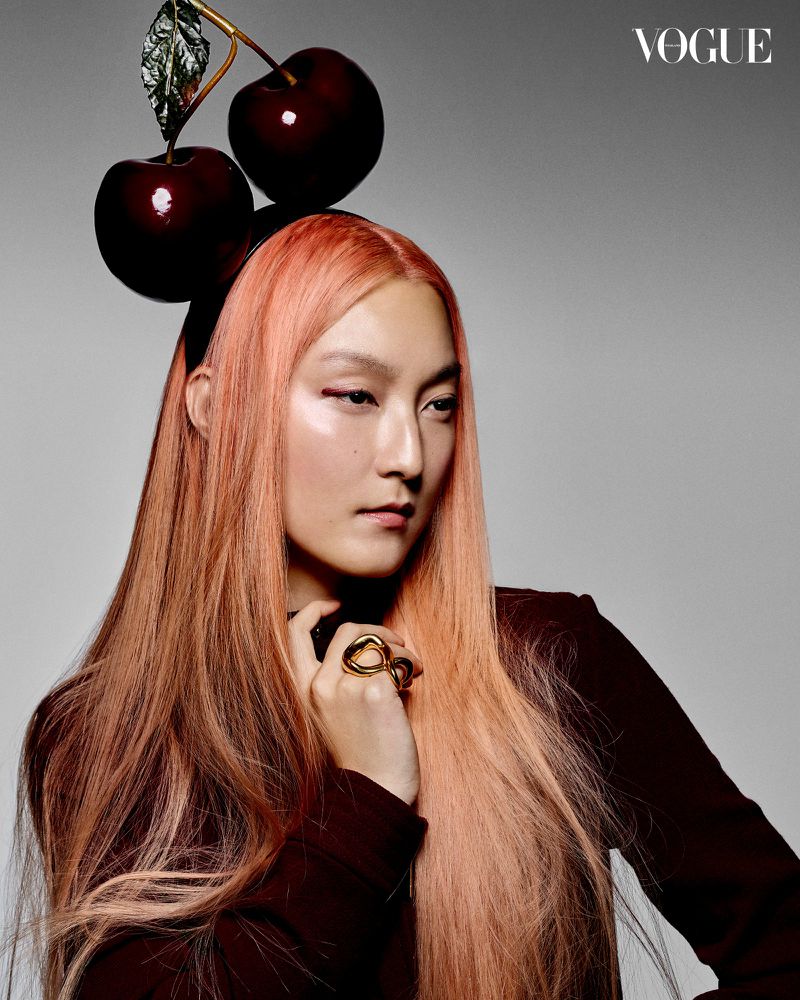 A fashion-style portrait of an Asian adult female with peach-colored hair, wearing a cherry-shaped headband, and striking a pose against a grey backdrop.