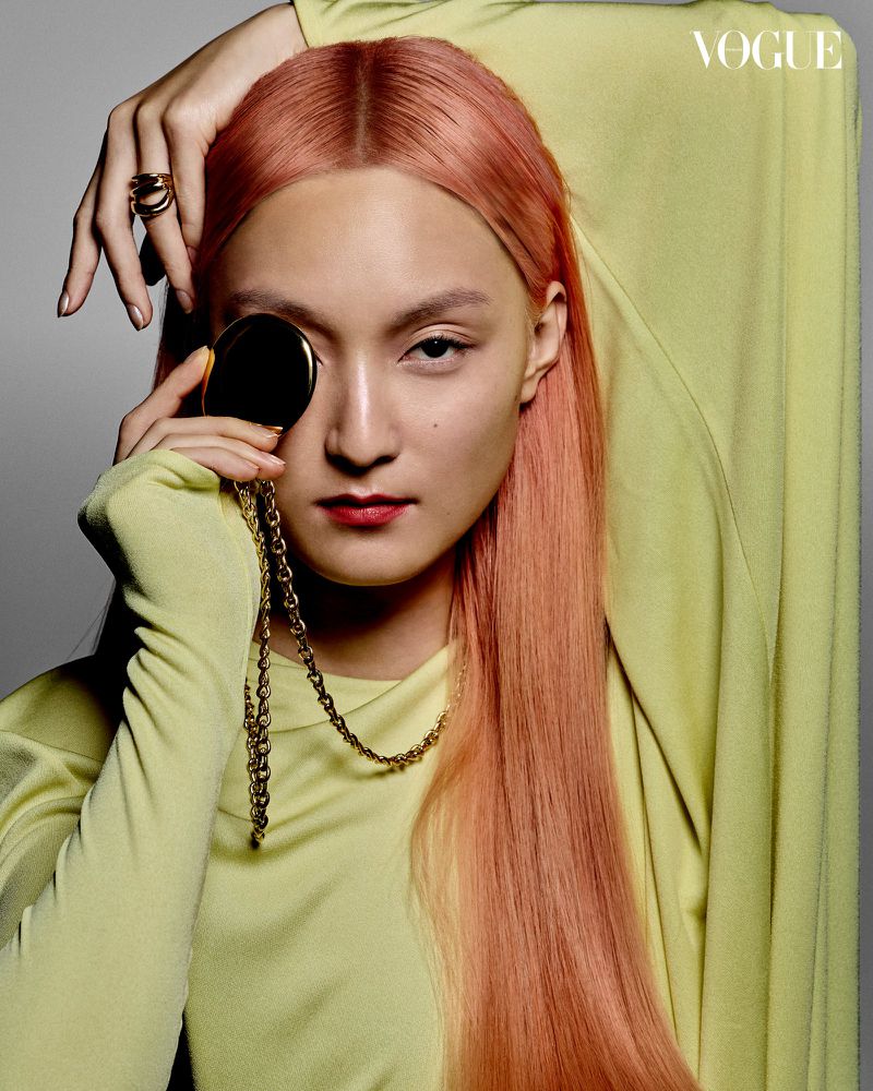 Close-up portrait of a young Asian woman with peach-colored hair, wearing a yellow top, holding a gold compact and chain.