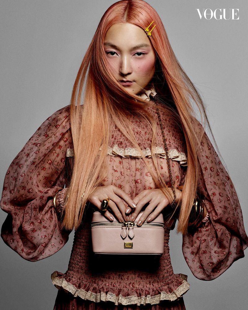 A fashionable Asian woman with orange-pink hair poses in a studio, wearing a floral dress and jewelry, holding a pink handbag.
