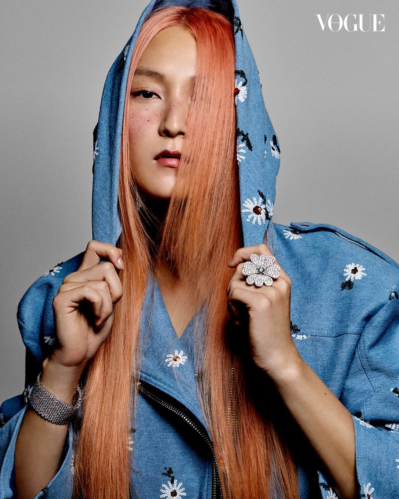 Close-up portrait of a fair-skinned person with peach-colored hair wearing a blue hooded jacket with flower designs against a grey background.