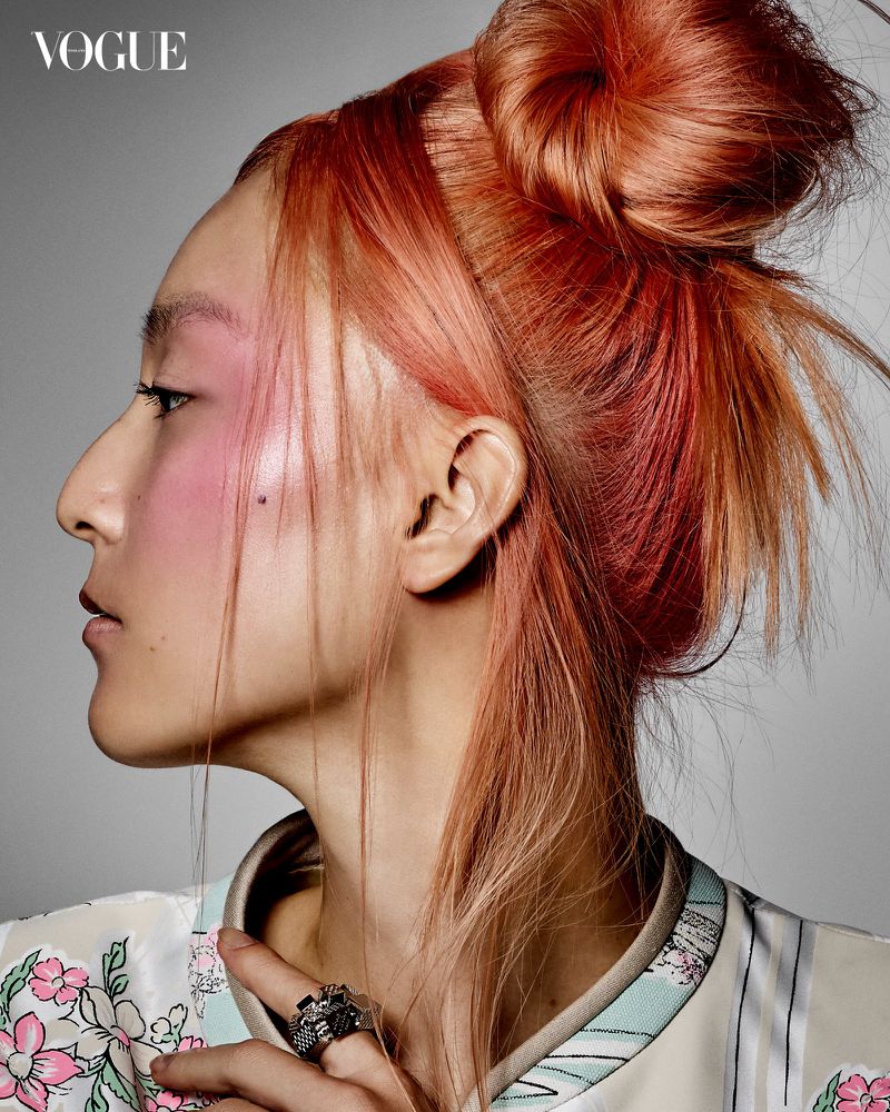 Portrait of a young woman with pink and orange hair, styled in a bun, wearing blush and a patterned top, against a gray background.