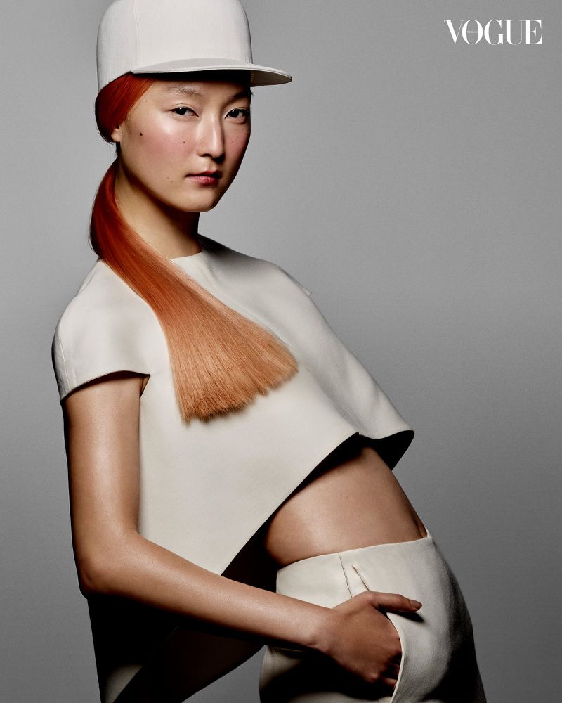 Fashion portrait of an Asian woman with orange hair, wearing a white top and baseball cap against a grey background.