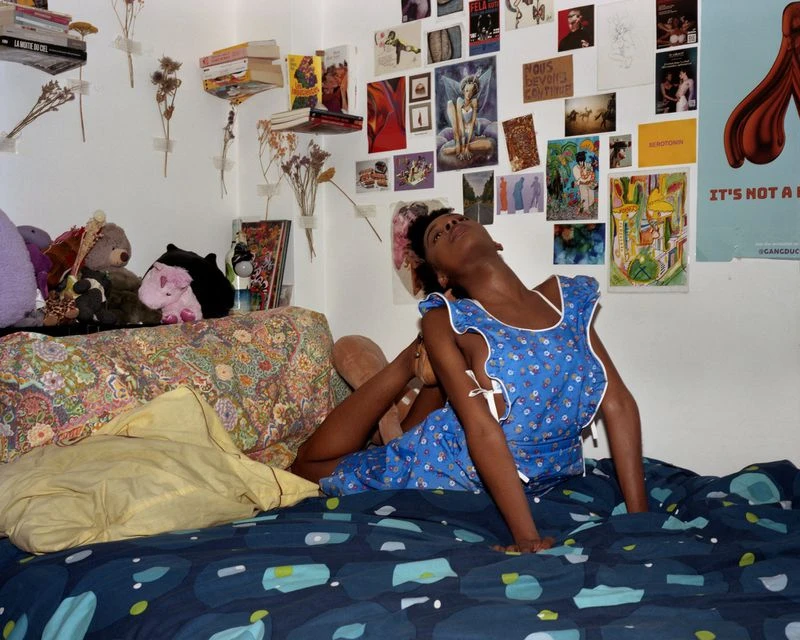 Young Black woman in blue floral dress sits on a bed, arching her back in an expressive pose, surrounded by eclectic room decor.